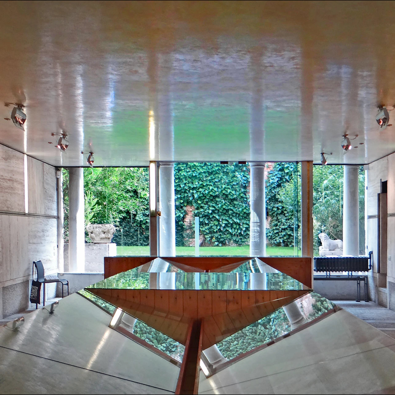 A modern room with reflective surfaces and a large window opening to a lush garden with trees and statues.