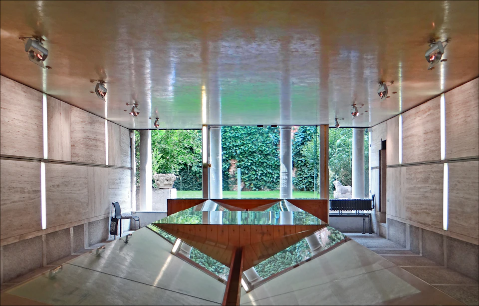 A modern room with reflective surfaces and a large window opening to a lush garden with trees and statues.