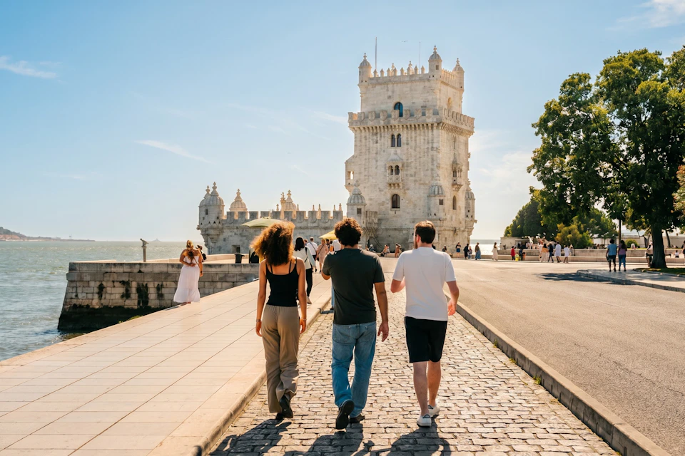 Three people walking on a cobblestone path toward a historic tower near water under a clear blue sky.