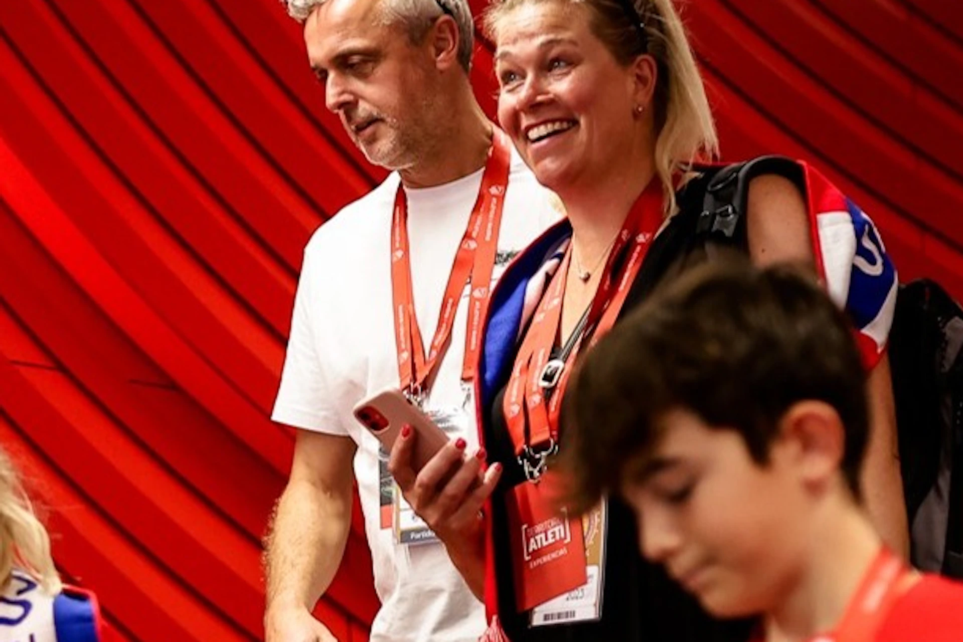A family in red and white football shirts and scarves descends stairs inside a stadium with a red wall background.