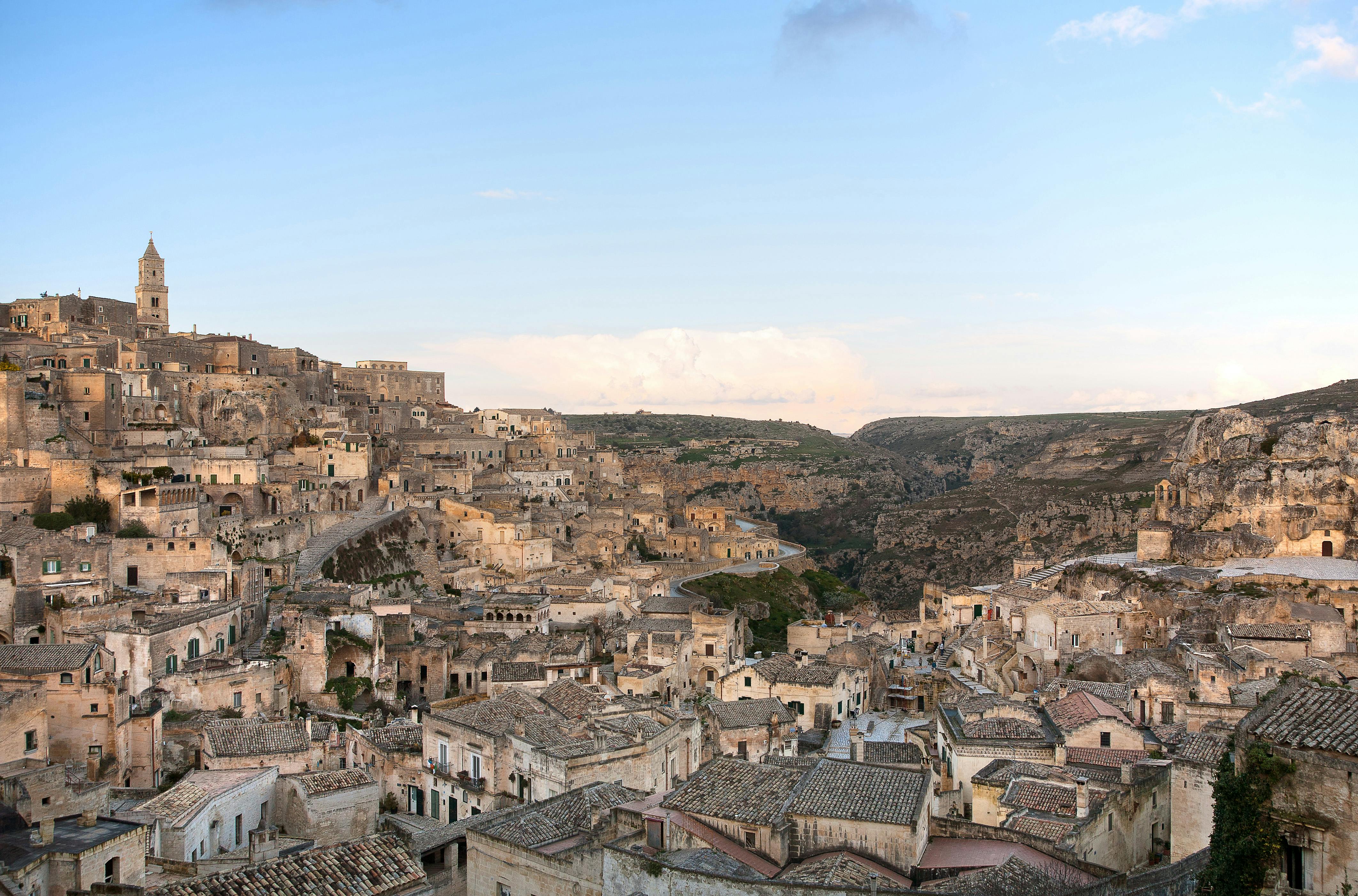 A panoramic view of an old town with stone buildings and tiled roofs built into a rocky hillside under a clear blue sky.