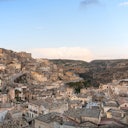A panoramic view of an old town with stone buildings and tiled roofs built into a rocky hillside under a clear blue sky.