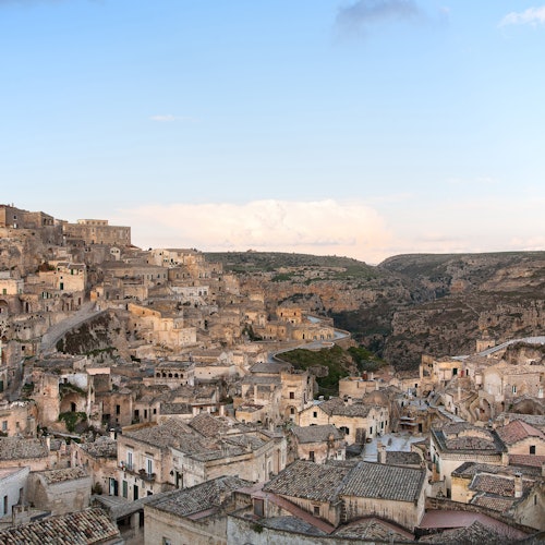 A panoramic view of an old town with stone buildings and tiled roofs built into a rocky hillside under a clear blue sky.