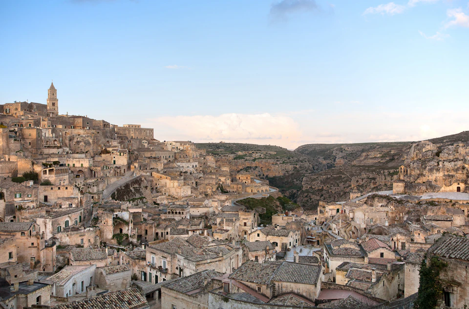 A panoramic view of an old town with stone buildings and tiled roofs built into a rocky hillside under a clear blue sky.
