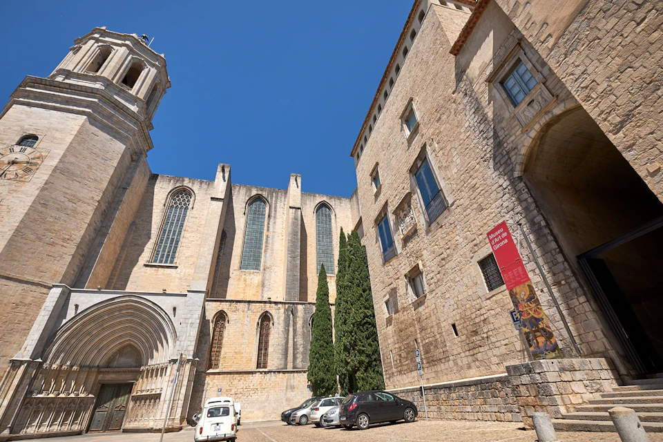 A historic stone building with arched windows and three parked cars under a clear blue sky. A red sign is attached to the building.