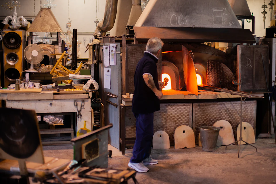 A man stands in front of a glass furnace in a workshop, holding a glassblowing pipe with molten glass. Tools and equipment surround him.