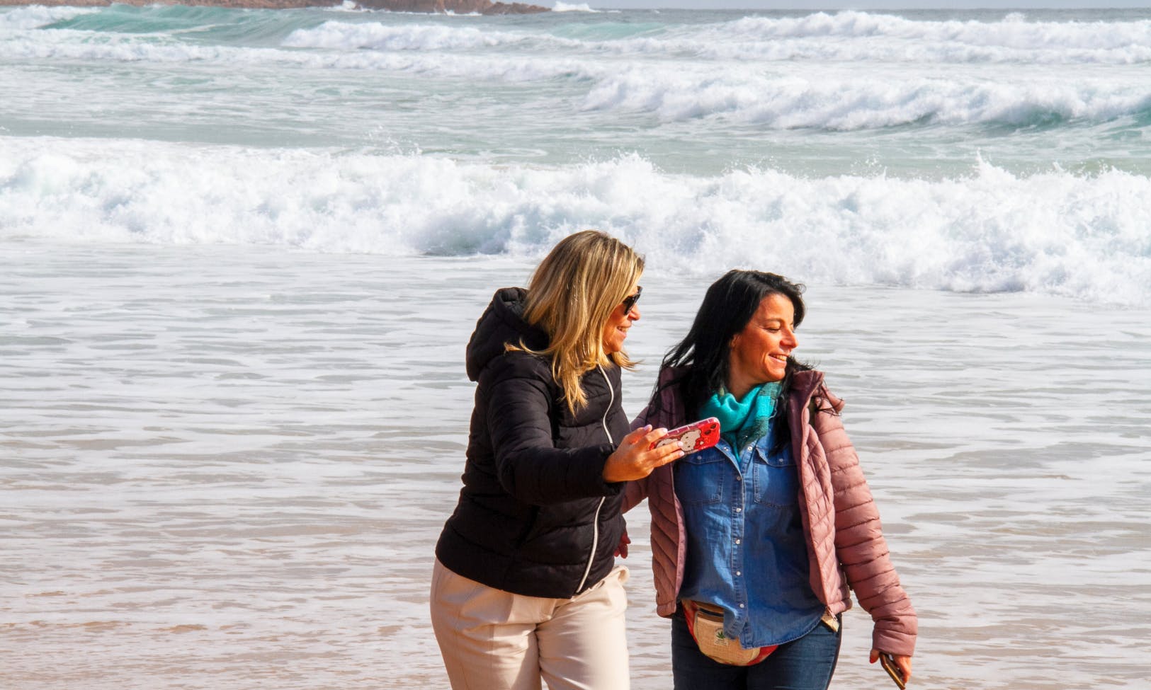 两位女士微笑着走在海滩上，后面海浪汹涌。 一位女士正在自拍。