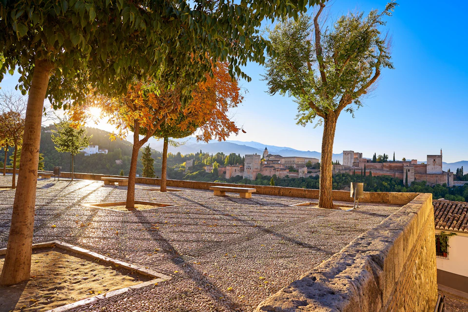 Sunlit courtyard with trees and benches, overlooking a historic fortress set against a backdrop of mountains and a clear blue sky.
