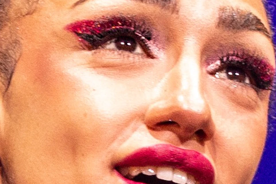A performer on stage singing into a microphone, wearing a studded outfit and a choker, with a vibrant purple-lit background.