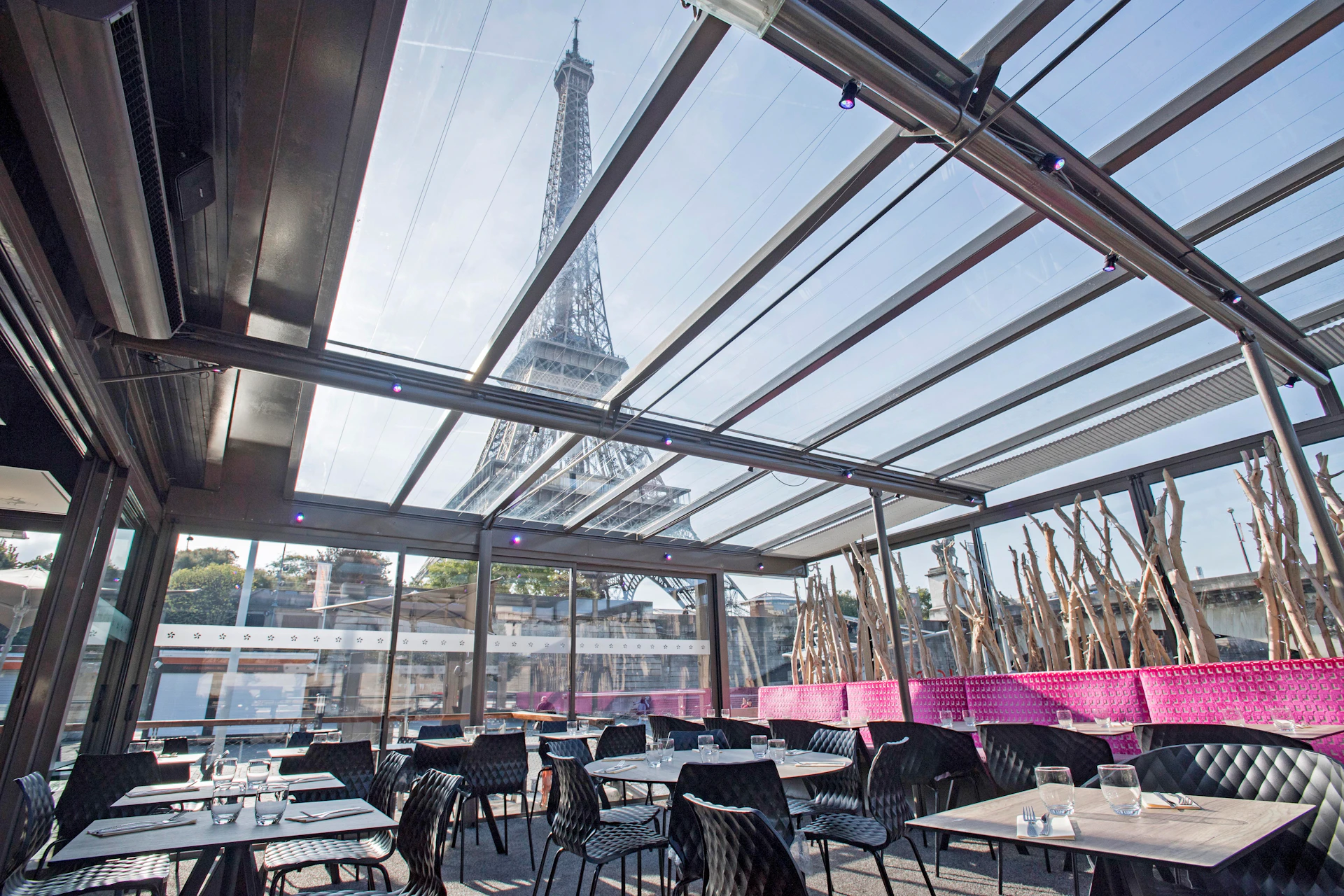 Outdoor café with glass roof offering a direct view of the Eiffel Tower, featuring black chairs and tables with purple seat cushions.