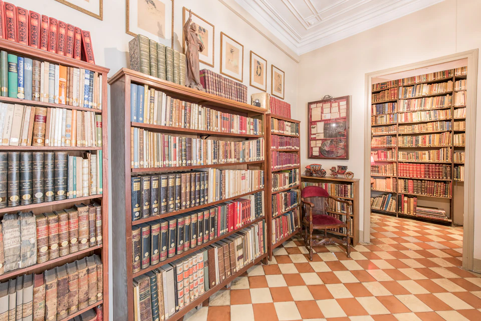 A cozy library with bookshelves filled with books, a wooden chair and small table, and framed pictures on the walls.