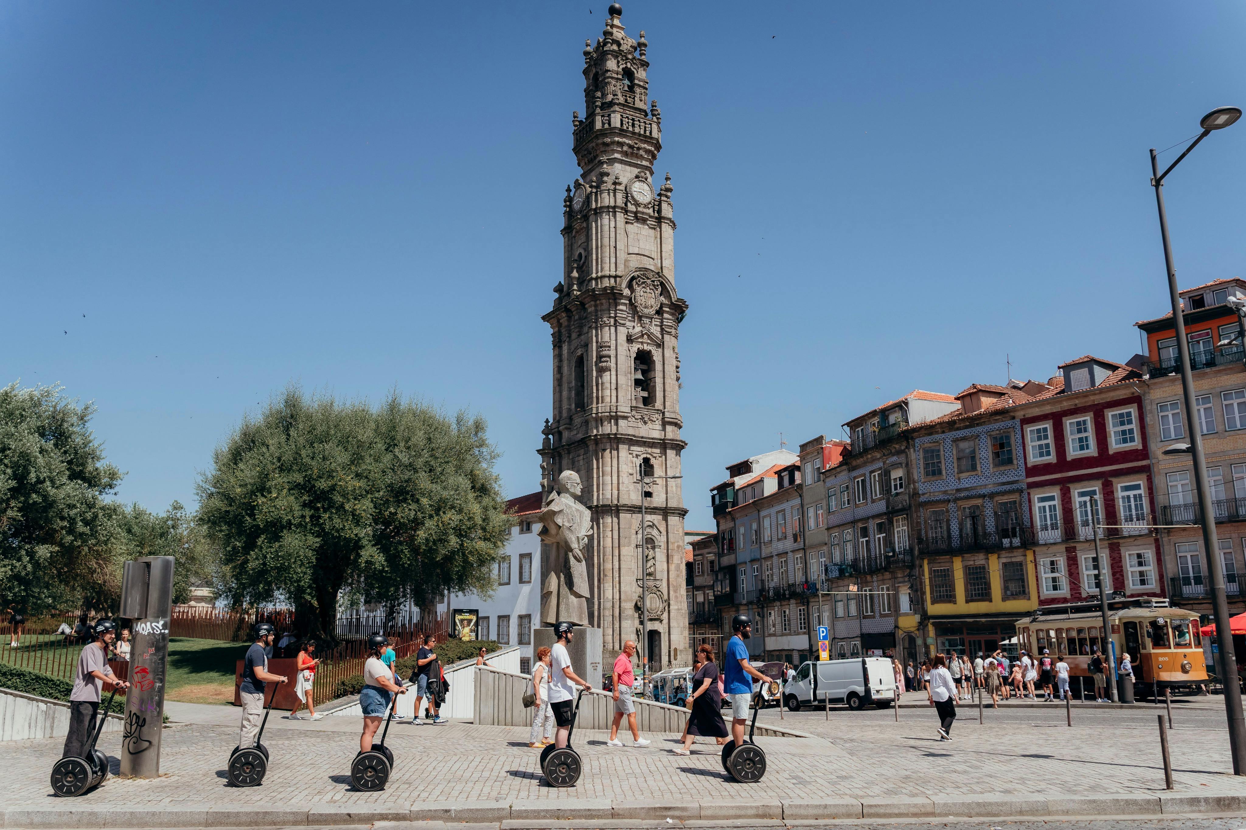 사람들은 오래된 건물로 둘러싸인 유럽 도시 광장의 높은 유서 깊은 시계탑과 동상 근처에서 세그웨이를 타고 있습니다.