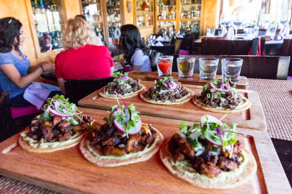 Wooden boards with tacos topped with greens and radishes on a restaurant table with drinks. Two people are seated in the background.