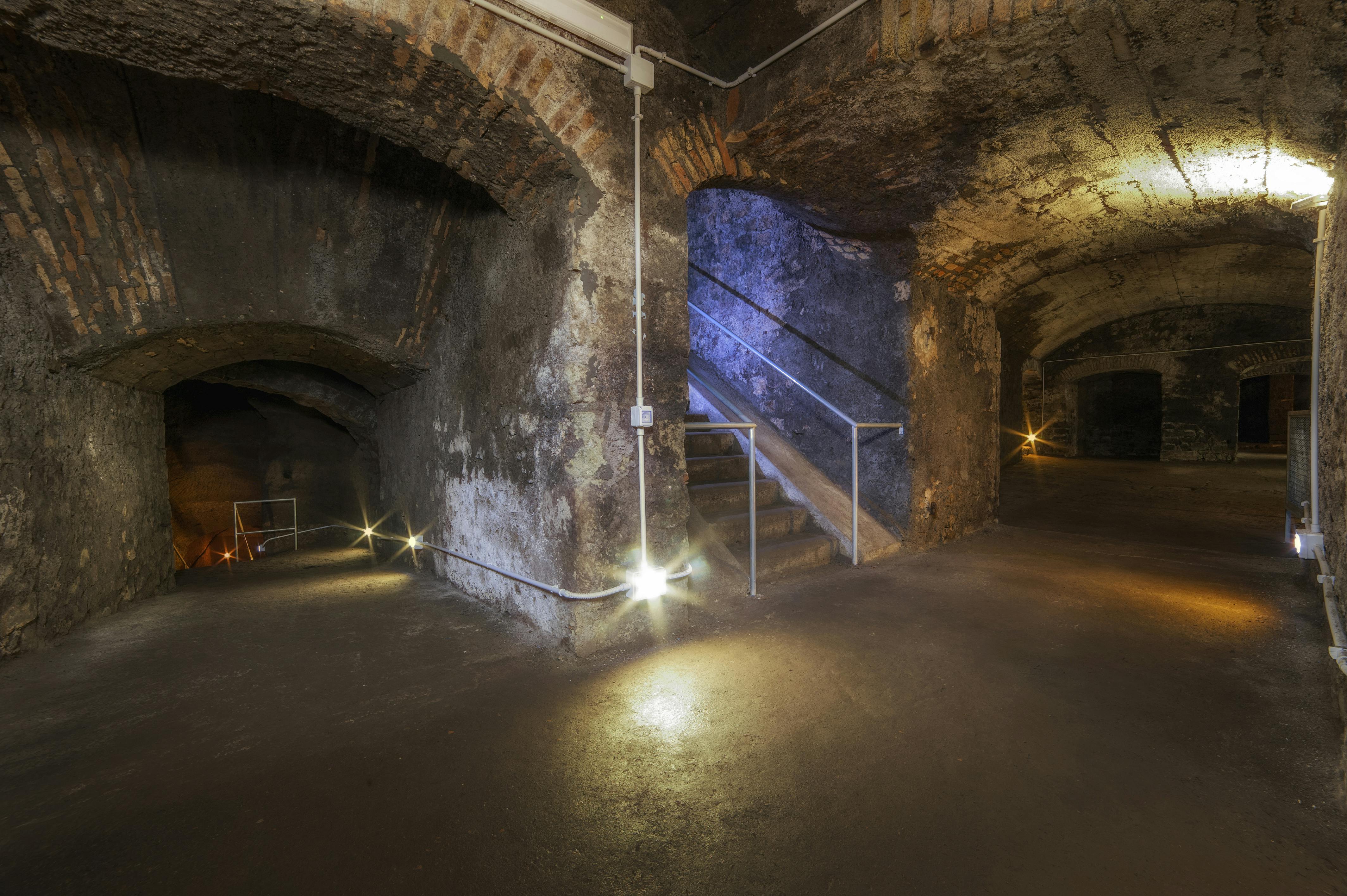 Svagt oplyst underjordisk kælder med stenmure, buede døråbninger og metalskinnede trapper oplyst af små lys.