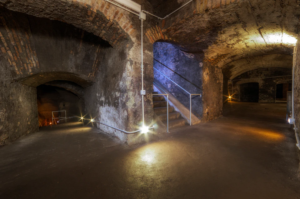 Dimly lit underground cellar with stone walls, arched doorways, and metal-railed stairs illuminated by small lights.