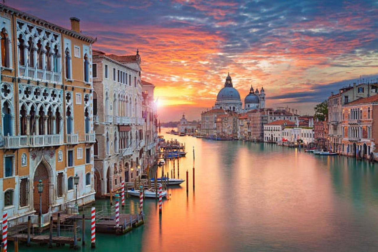 A canal in Venice at sunset, with historic buildings on both sides, gondolas docked, and a church with domes in the distance.