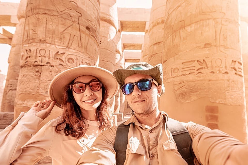Two people wearing hats and sunglasses smile for a selfie in front of ancient stone pillars with hieroglyphics.