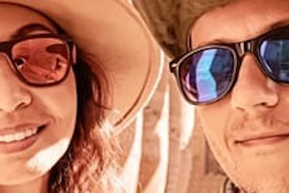 Two people wearing hats and sunglasses smile for a selfie in front of ancient stone pillars with hieroglyphics.