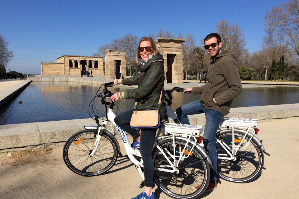 Two people on bicycles pose in front of the Debod Temple, with a reflecting pool and clear blue sky.