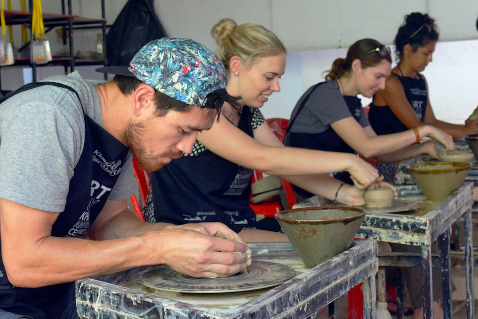 A group of people engaged in pottery-making, shaping clay on spinning wheels, seated in a workshop setting.
