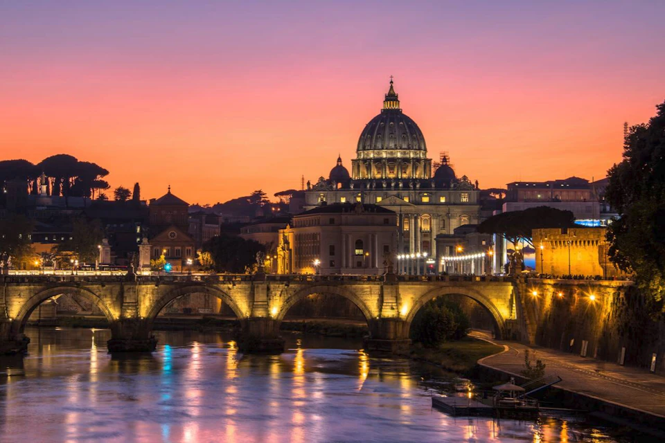 Illuminated bridge and St. Peter's Basilica in Rome at sunset, with a colorful sky and reflections on the water.