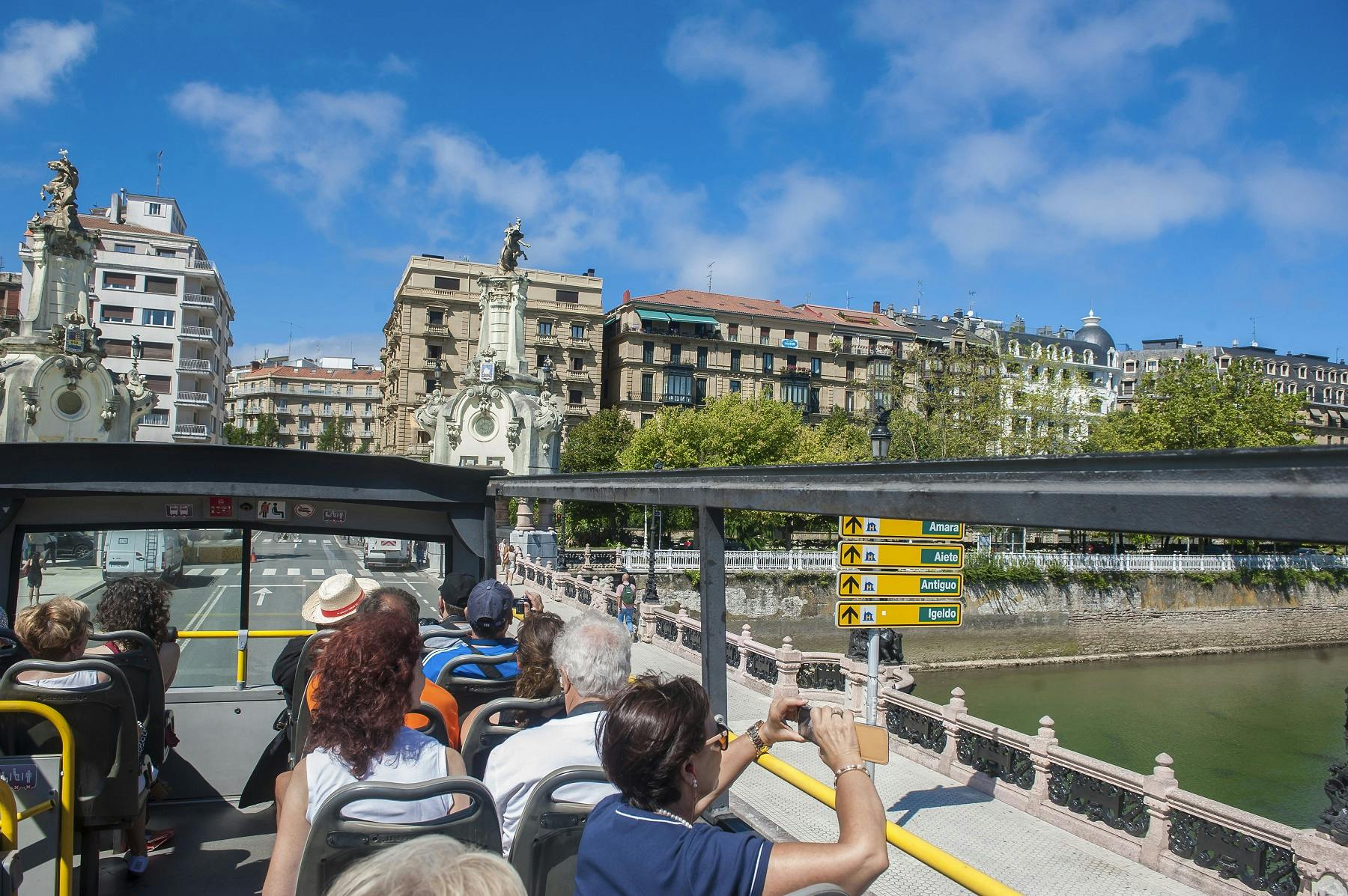 A red double-decker tour bus with "Donostia San Sebastián" on the side, filled with passengers, drives along a coastal road.
