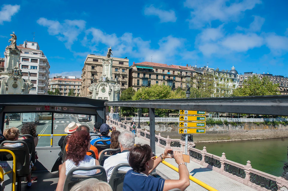 A red double-decker tour bus with "Donostia San Sebastián" on the side, filled with passengers, drives along a coastal road.