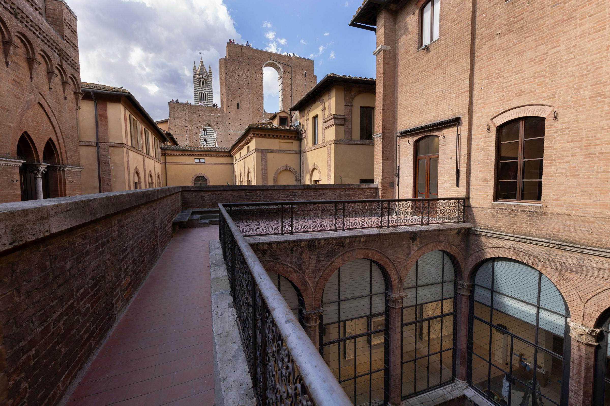 Cortile storico con finestre ad arco, ringhiere intricate ed edifici gialli e marroni. Un muro in rovina è visibile sullo sfondo.
