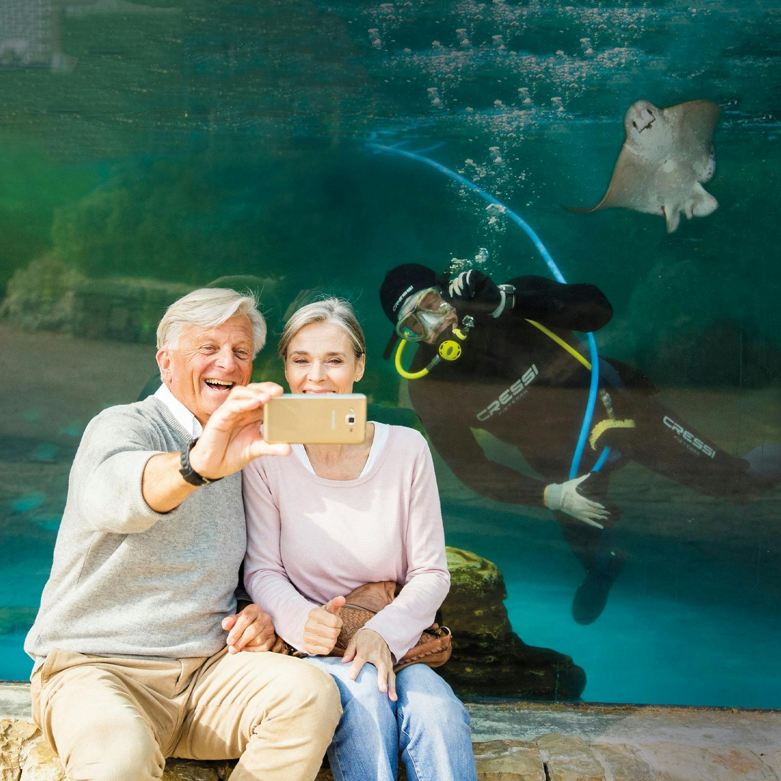 An elderly couple takes a selfie in front of an aquarium, with a diver and a stingray visible in the background.