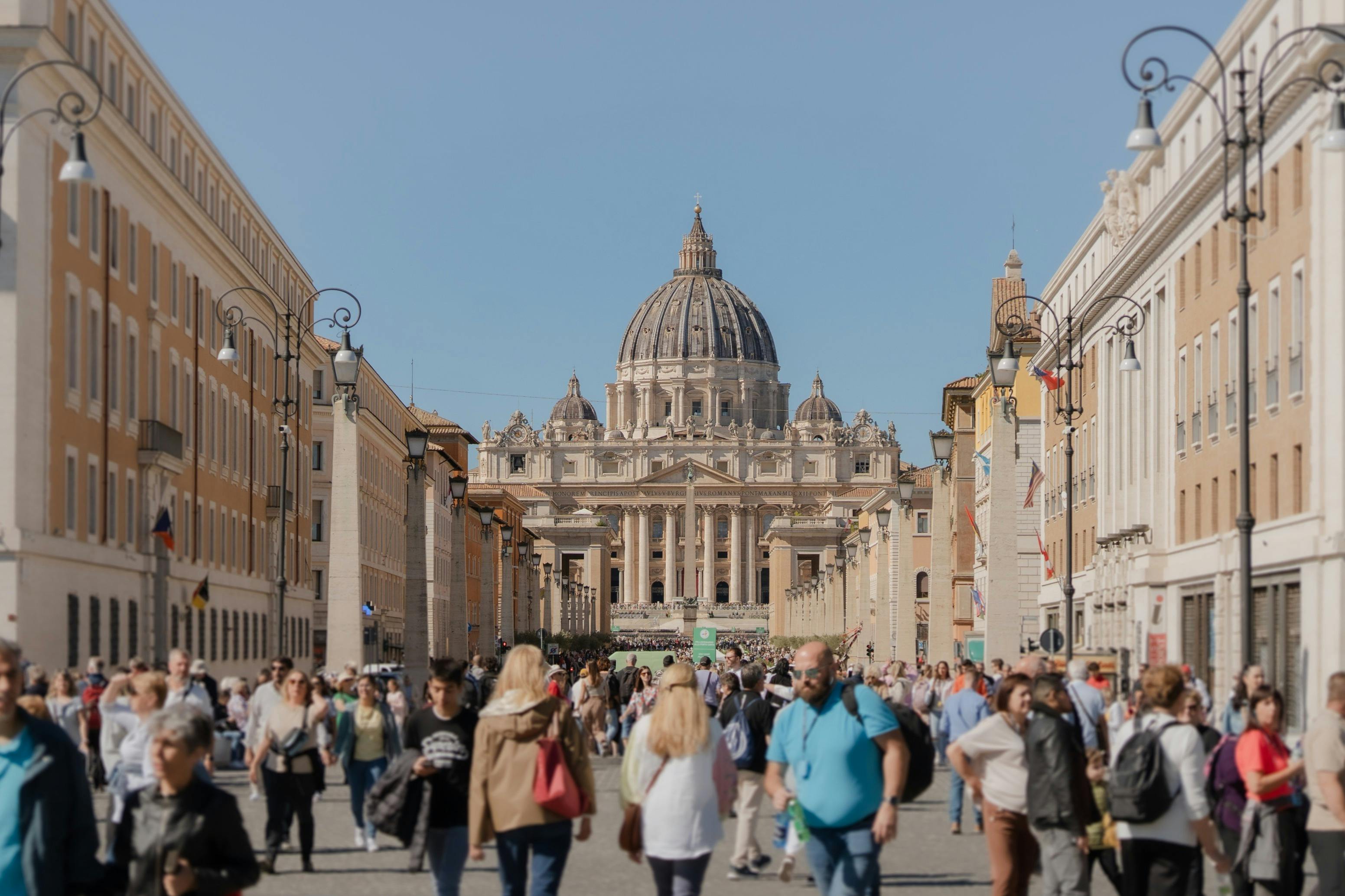 一条繁忙的街道，游客走向一座遥远的大型圆顶建筑，两侧是晴朗的蓝天下的历史建筑。
