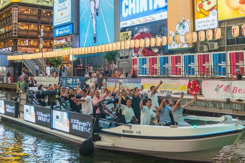 A boat filled with cheering people passes under illuminated lanterns in a busy city area with colorful billboards.