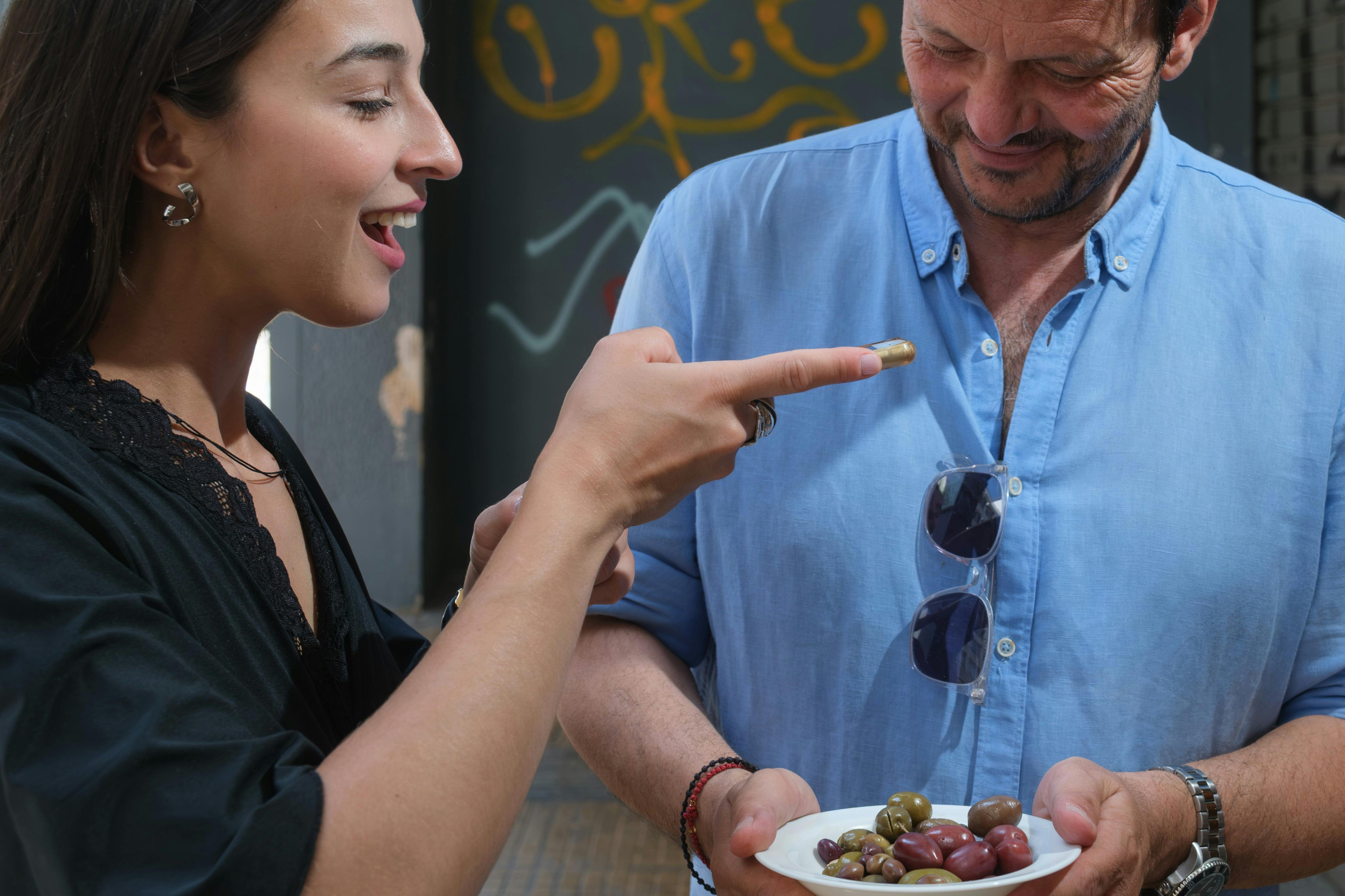 Μια γυναίκα και ένας άντρας μοιράζονται ελιές. Δείχνει παιχνιδιάρικα με μία στο δάχτυλό της. Ο άντρας κρατάει ένα άσπρο πιάτο με ελιές.