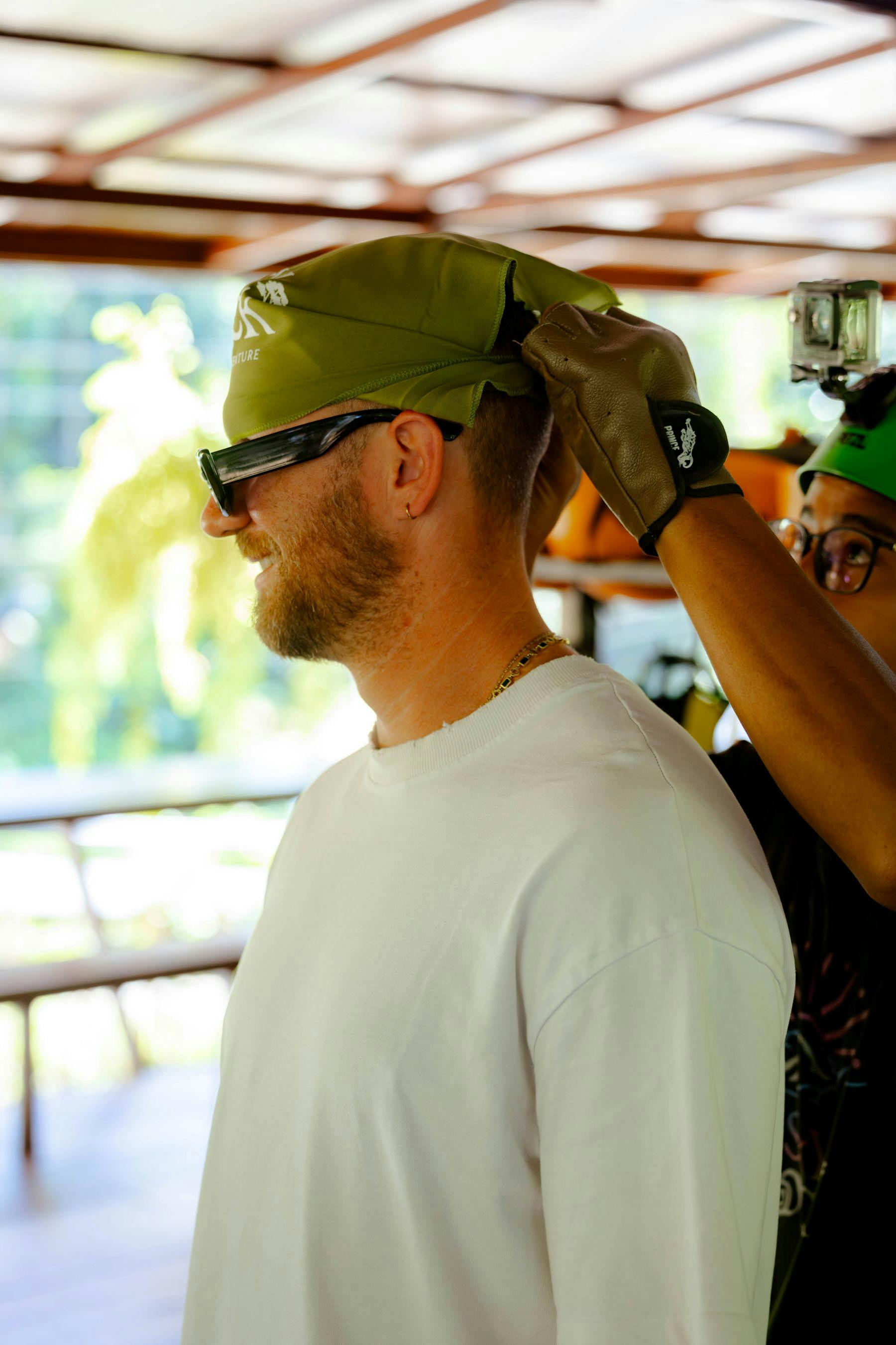 A person in a white shirt is having a green bandana adjusted on their head by another person wearing gloves and glasses.