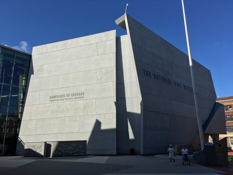 The exterior of The National WWII Museum with large concrete walls, shadows, and a few visitors walking nearby.