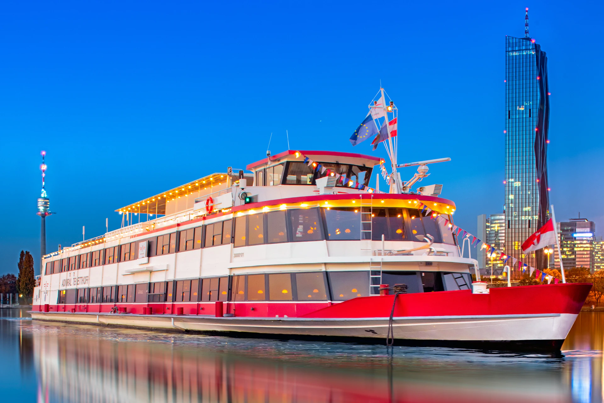 A white and red boat decorated with string lights and flags, docked at a harbor against a clear blue evening sky.