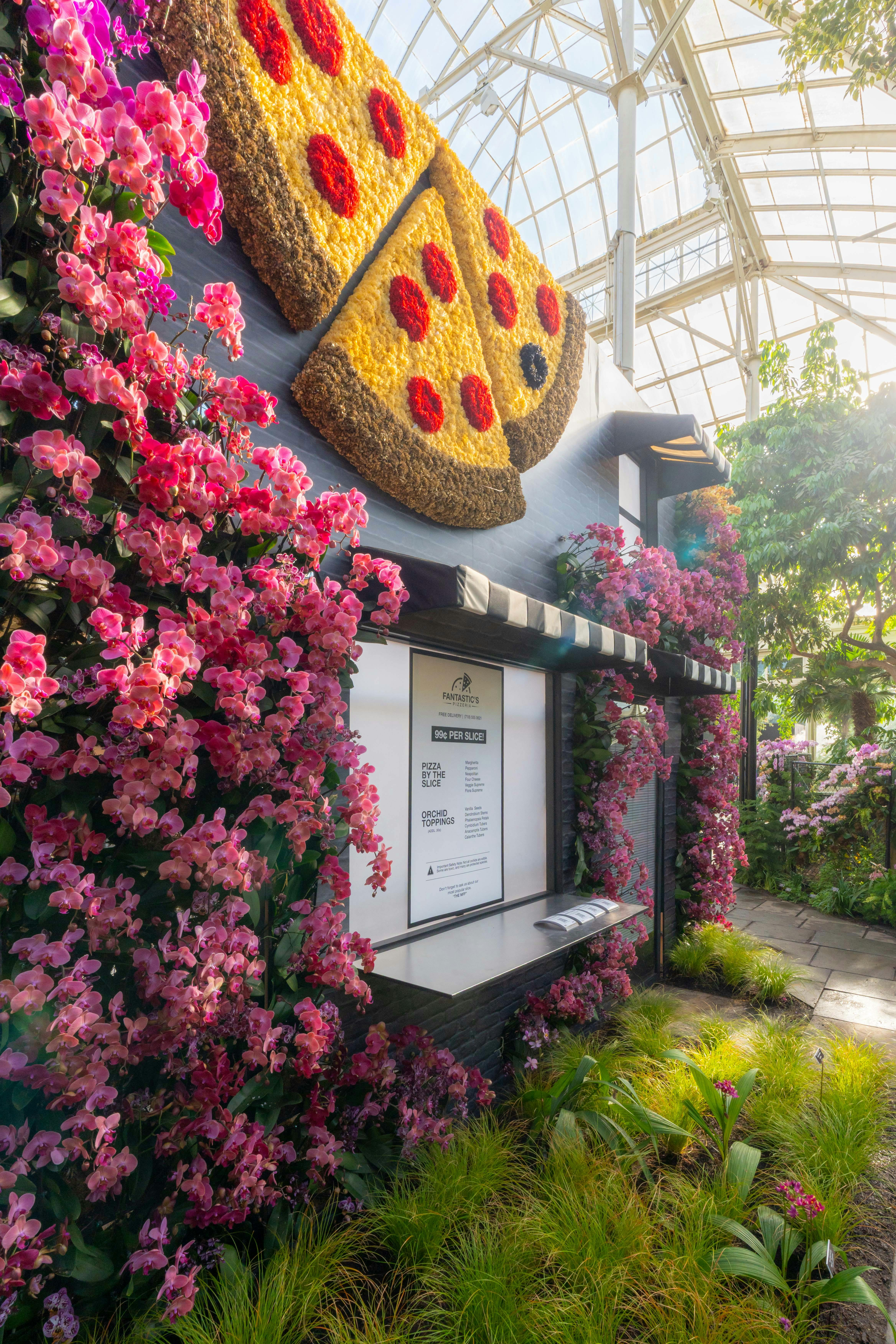Una mostra de jardí amb un edifici adornat amb flors roses i una gran decoració de rodanxes de pizza a la paret.