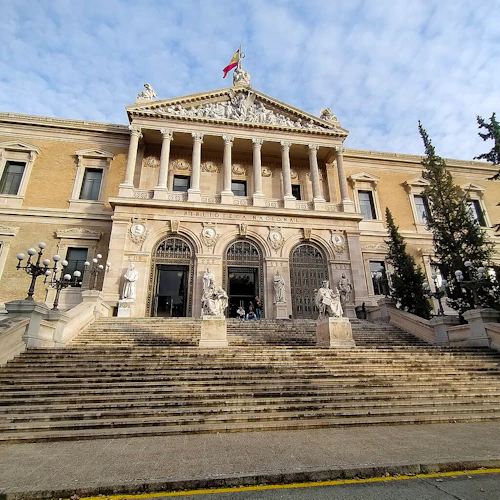Stor stentrappa som leder till Biblioteca Nacional-byggnaden med pelare, statyer och nationell flagga ovanpå under en delvis molnig himmel.