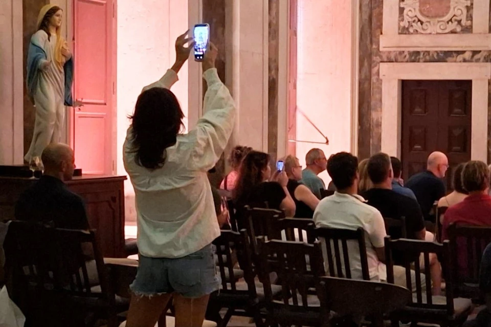A woman in casual clothing stands, holding up a smartphone to take a photo in a room with seated people and classical decor.