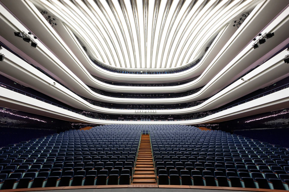 Modern theater interior with rows of blue seats, wooden center aisle, and multi-tiered balconies under futuristic, wave-like ceiling.