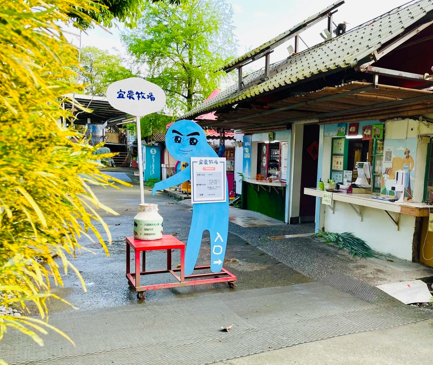An outdoor market area with a blue character cutout holding a sign near a small red table with a bottle, surrounded by greenery.