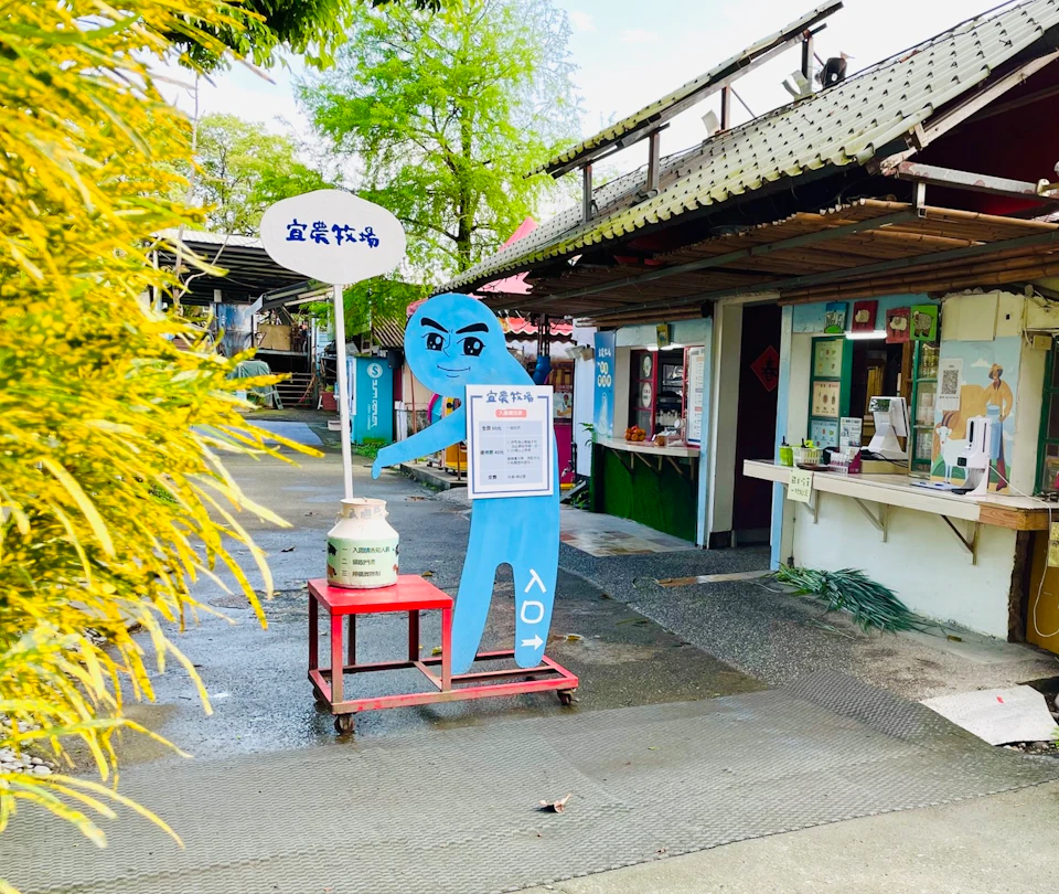 A street scene with a blue cartoon figure holding a sign, next to a cart with a milk can, in front of a small shop.