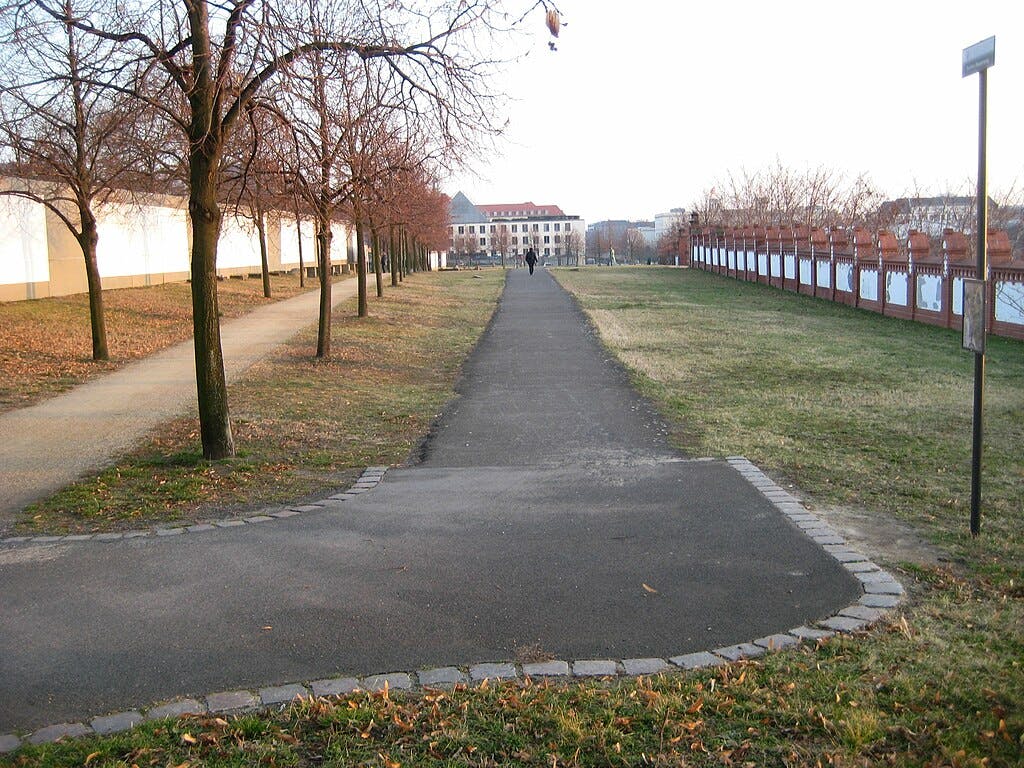Sentier bordé d'arbres aux feuilles clairsemées, menant à des bâtiments au loin. Peu de personnes marchent le long du chemin.