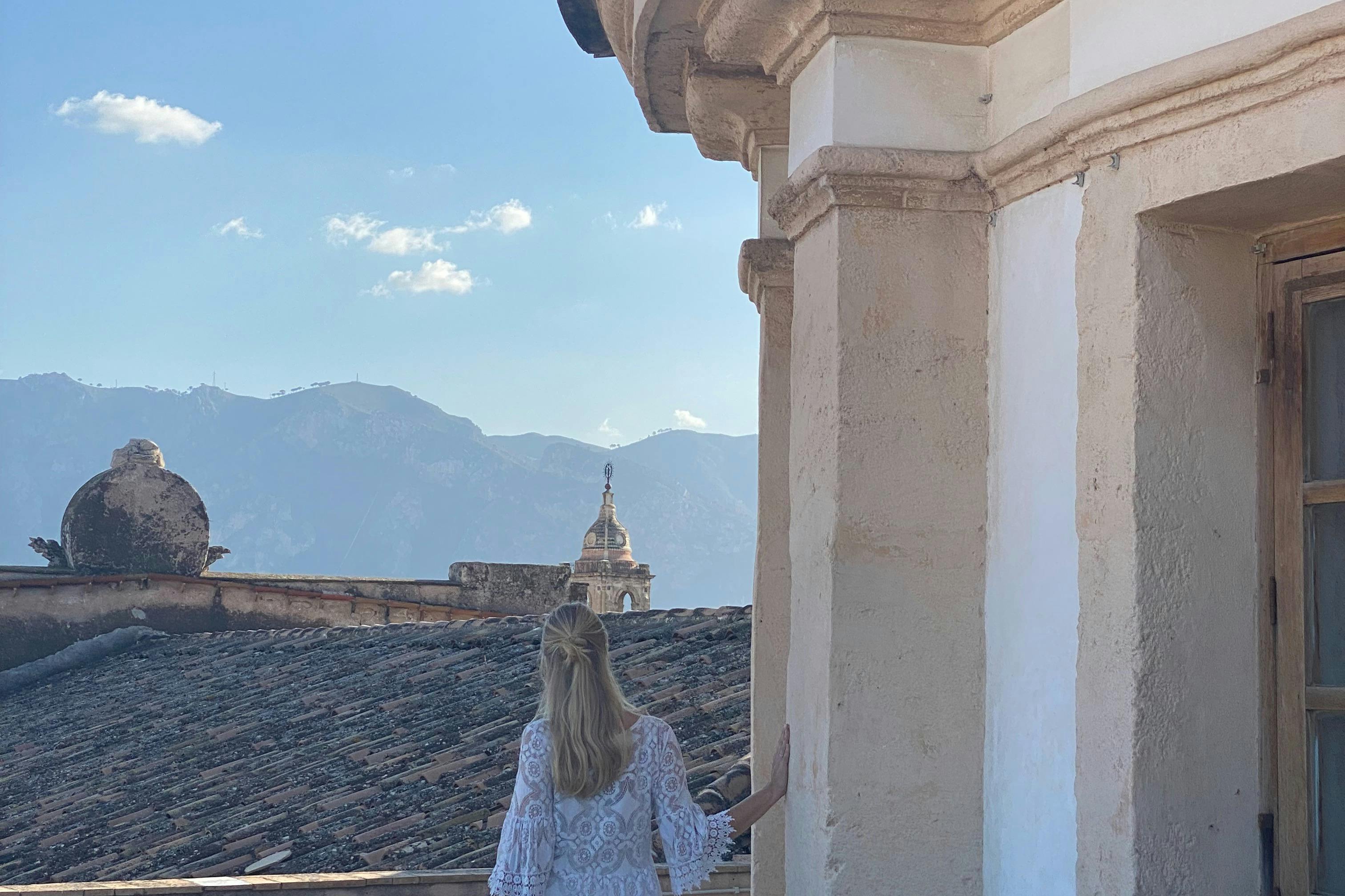 Une personne aux cheveux longs se tient près d'une structure en pierre, surplombant les toits et une chaîne de montagnes lointaine sous un ciel dégagé.