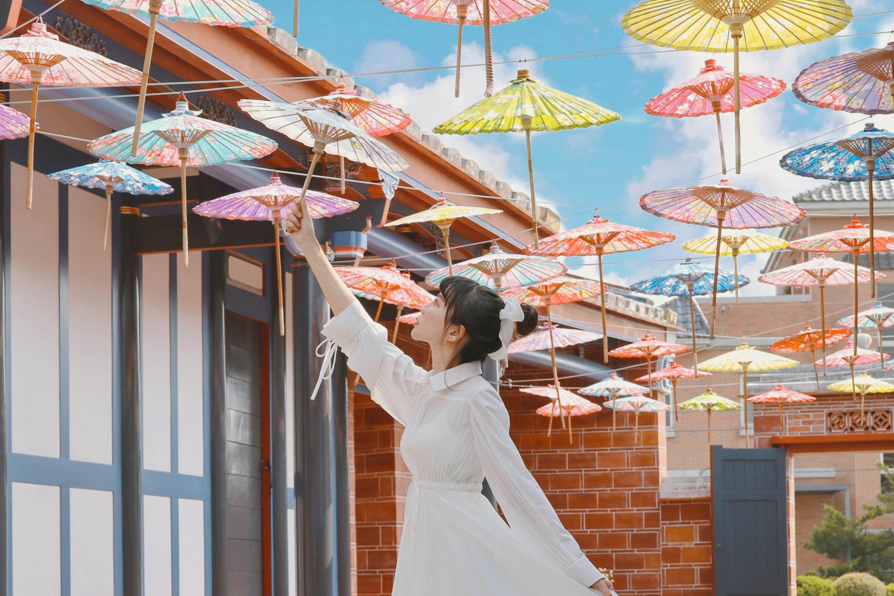 Person in white dress reaching for colorful paper umbrellas hanging in a sunny courtyard.
