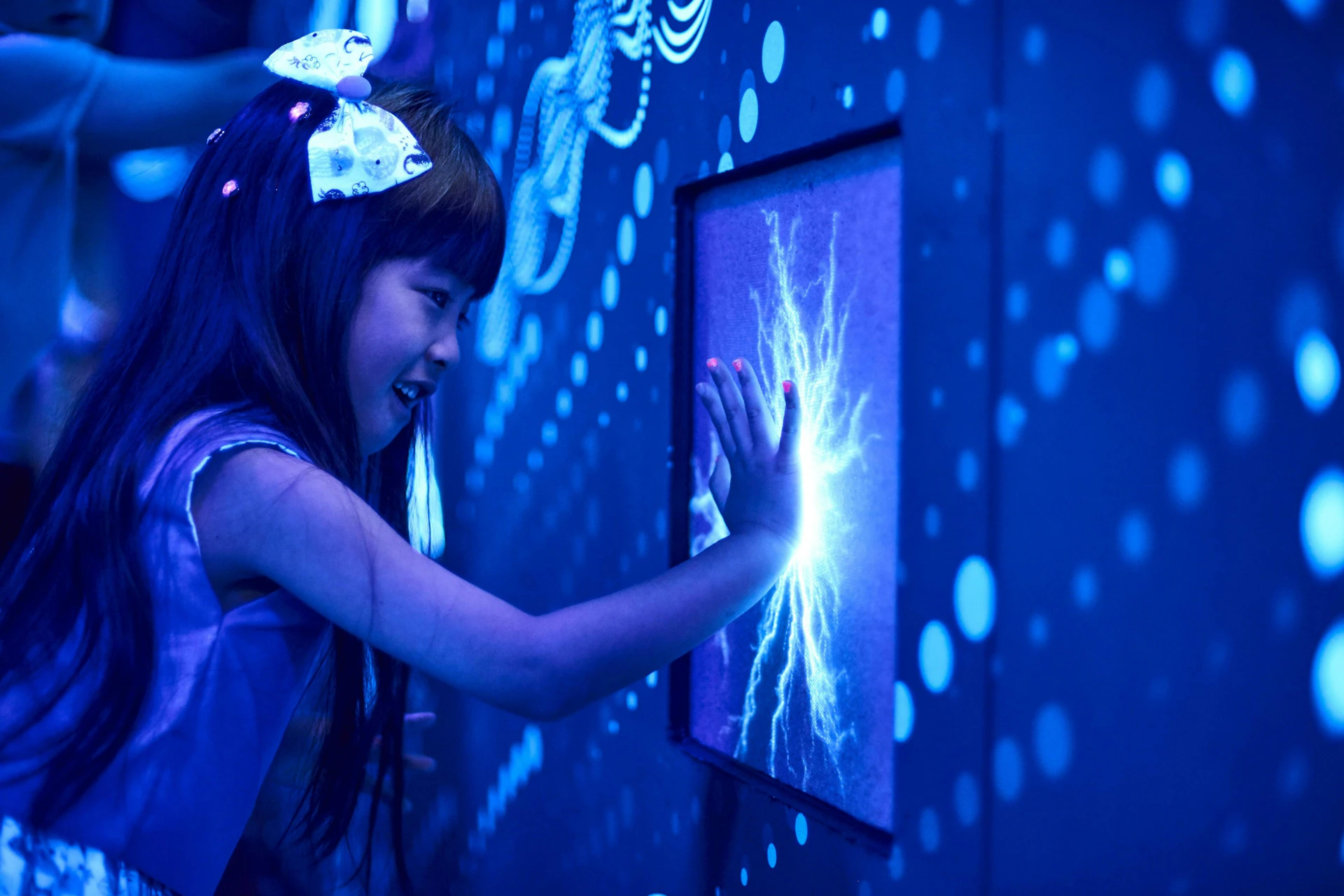 A child touches a glowing interactive display emitting blue light, surrounded by a high-tech, illuminated environment.