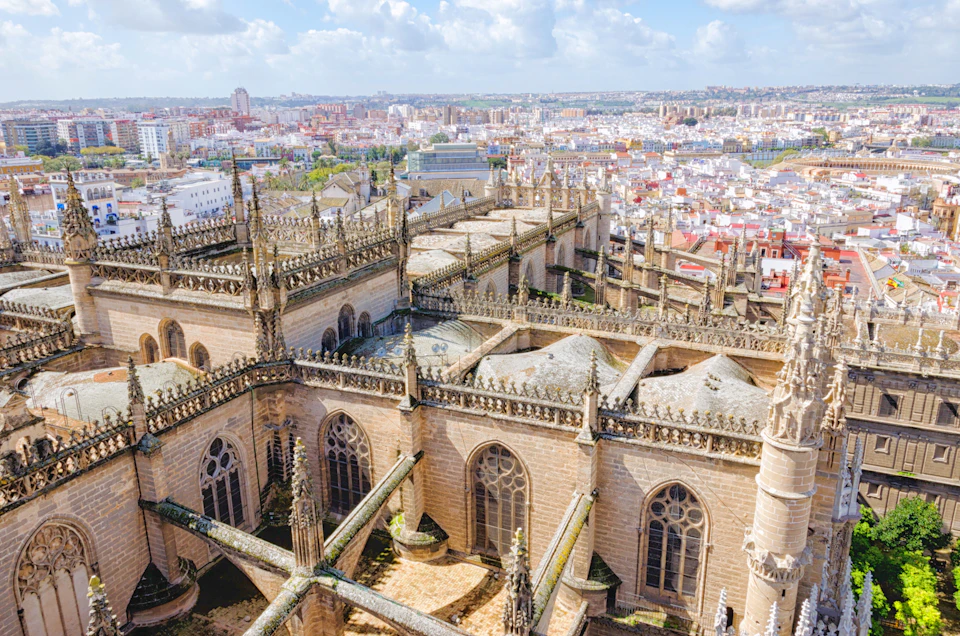 Aerial view of a large, ornate Gothic cathedral with spires and detailed carvings, set against a sprawling cityscape under a partly cloudy sky.