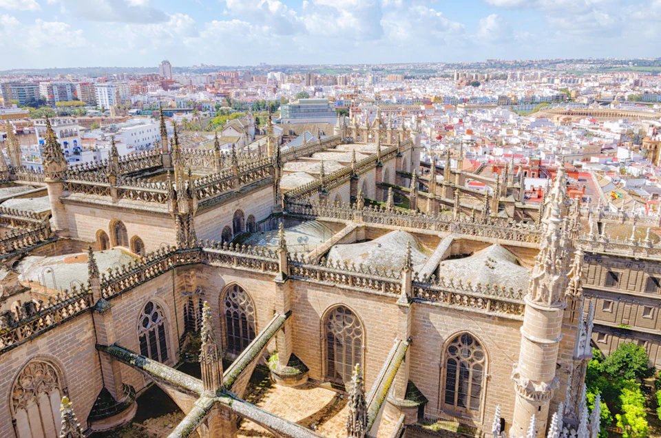 Aerial view of a large, ornate Gothic cathedral with spires and detailed carvings, set against a sprawling cityscape under a partly cloudy sky.