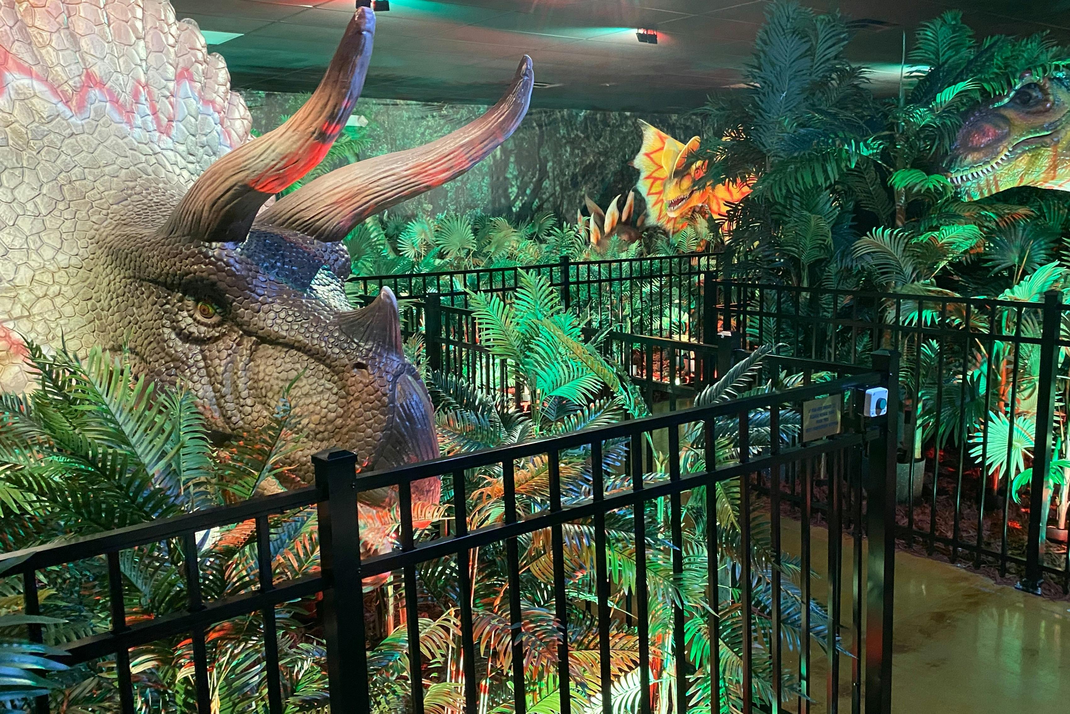 Exposición interior con modelos de dinosaurios a tamaño real entre una exuberante vegetación artificial, rodeada por una barandilla negra.