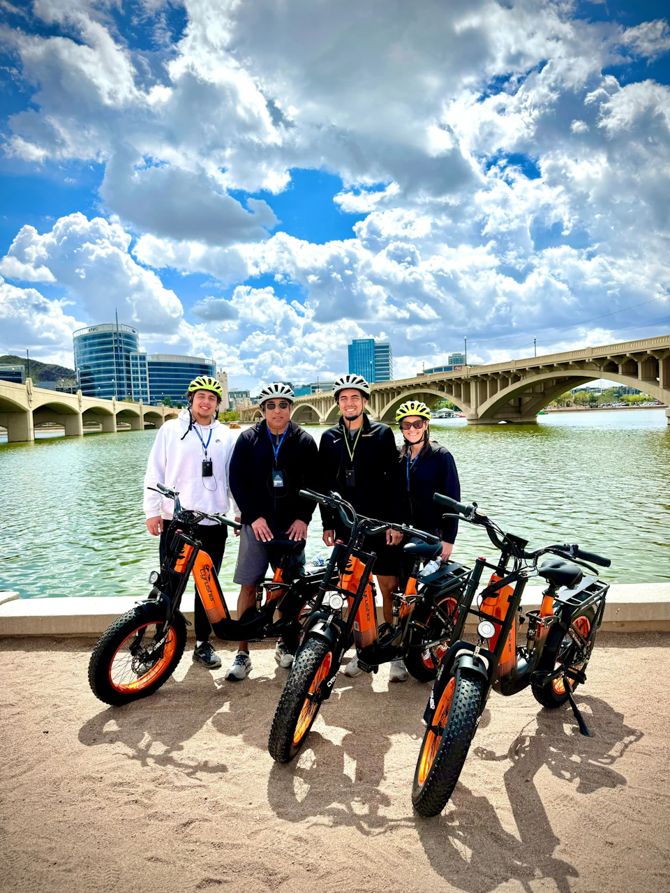 Four people wearing helmets stand behind electric bicycles by a waterfront, with a bridge and buildings in the background.