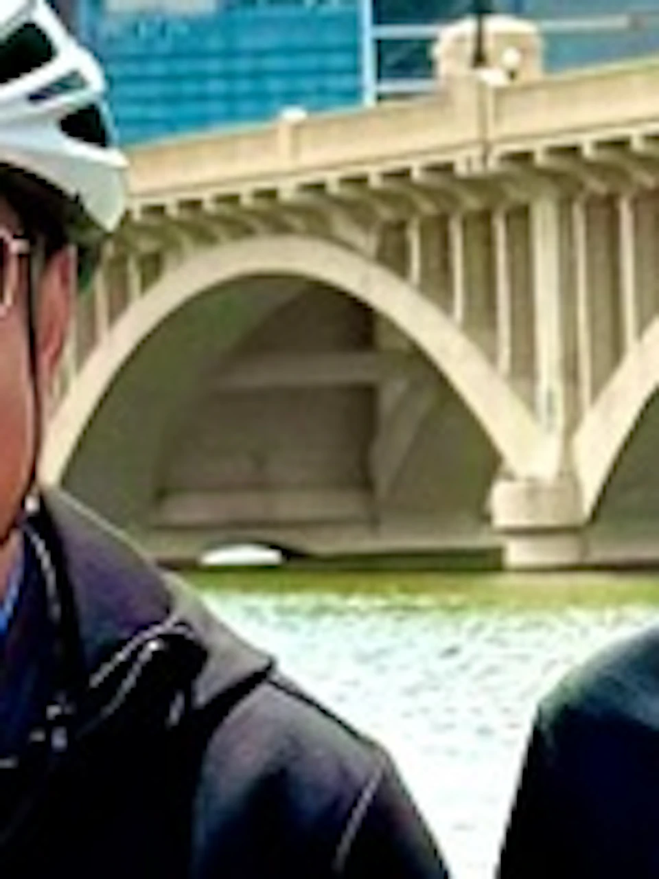 Four people wearing helmets stand behind electric bicycles by a waterfront, with a bridge and buildings in the background.